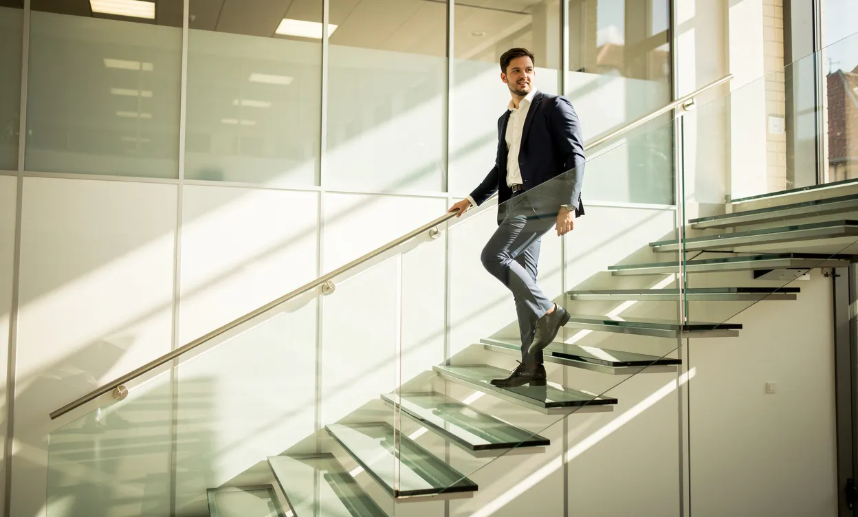 Un jeune professionnel monte un escalier en verre dans un bureau lumineux à Lille, baigné de soleil et d'ombres.